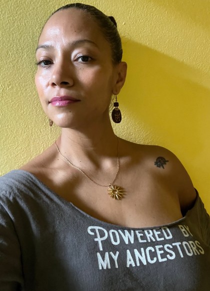 Photo of author Zayda Rivera standing in front of a yellow wall, looking at the camera, wearing a shirt that says “Powered By My Ancestors”